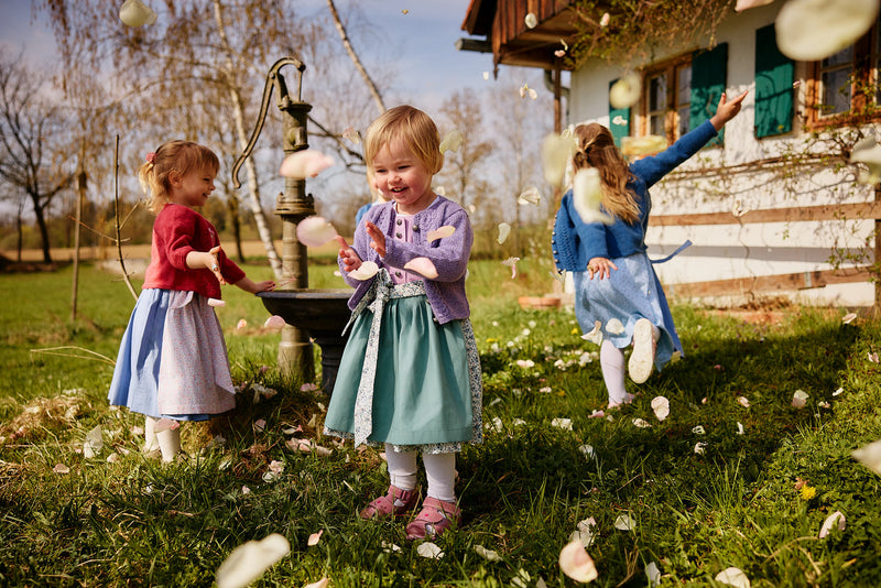 Farbenfrohe, unterschiedliche Kinderdirndl getragen von drei Kindern, die auf einer Wiese spielen