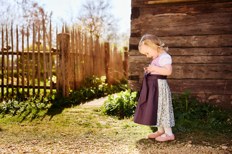 Kinderdirndl in flieder und lila mit blumigem Rock getragen von einem Mädchen vor einem Holzhaus, das das Dirndl bewundert