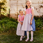Kinderdirndl in Altrosa mit blauem Rock und blumiger Schürze mit schönen weiß-rosafarbenen Trachtenknöpfen, getragen von zwei Mädchen in einem Garten.
