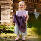 Kinderdirndlbluse und Kinderdirndl fliederfarben mit lila Schürze getragen von einem Kleinkind in einem Garten