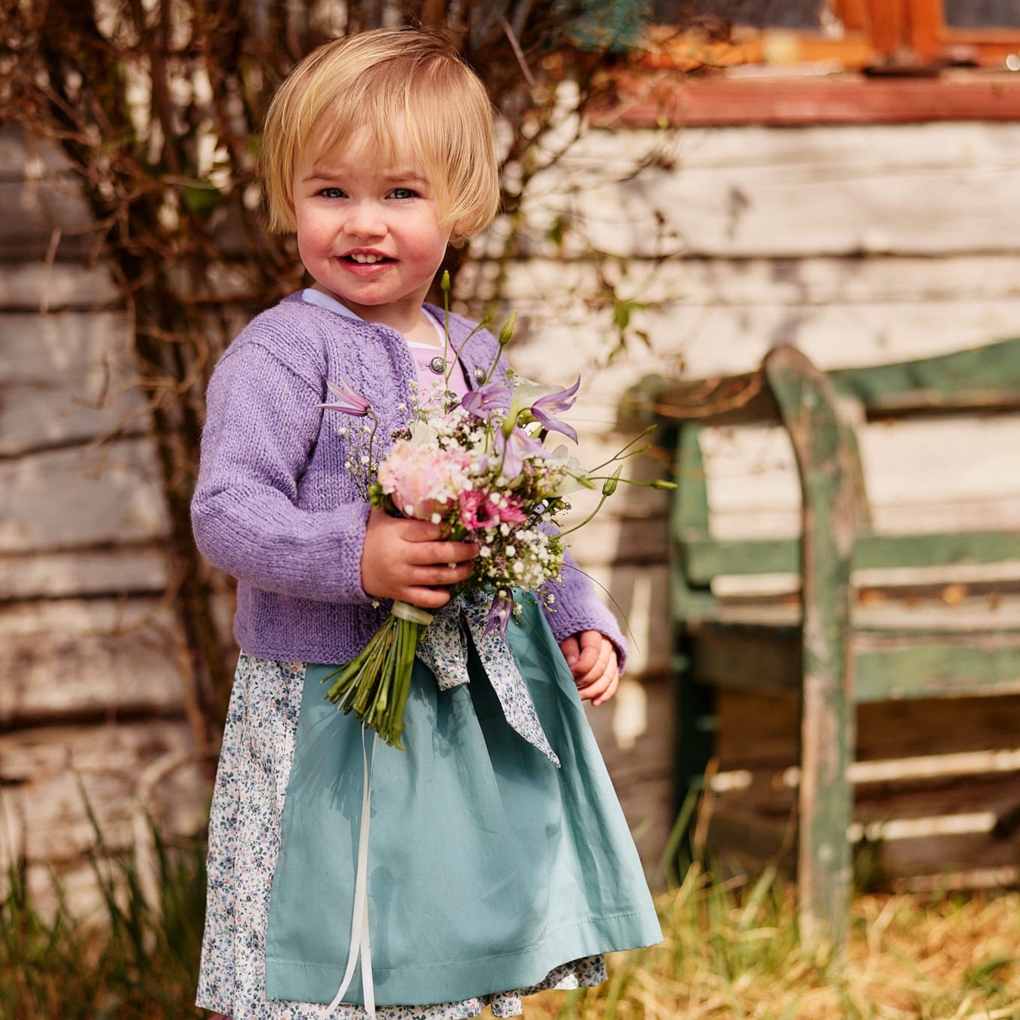 Kinderdirndl fliederfarben mit einer salbeifarbenen Schürze und einem blumigen Rock getragen von einem Kleinkind mit Blumenstrauß im Garten