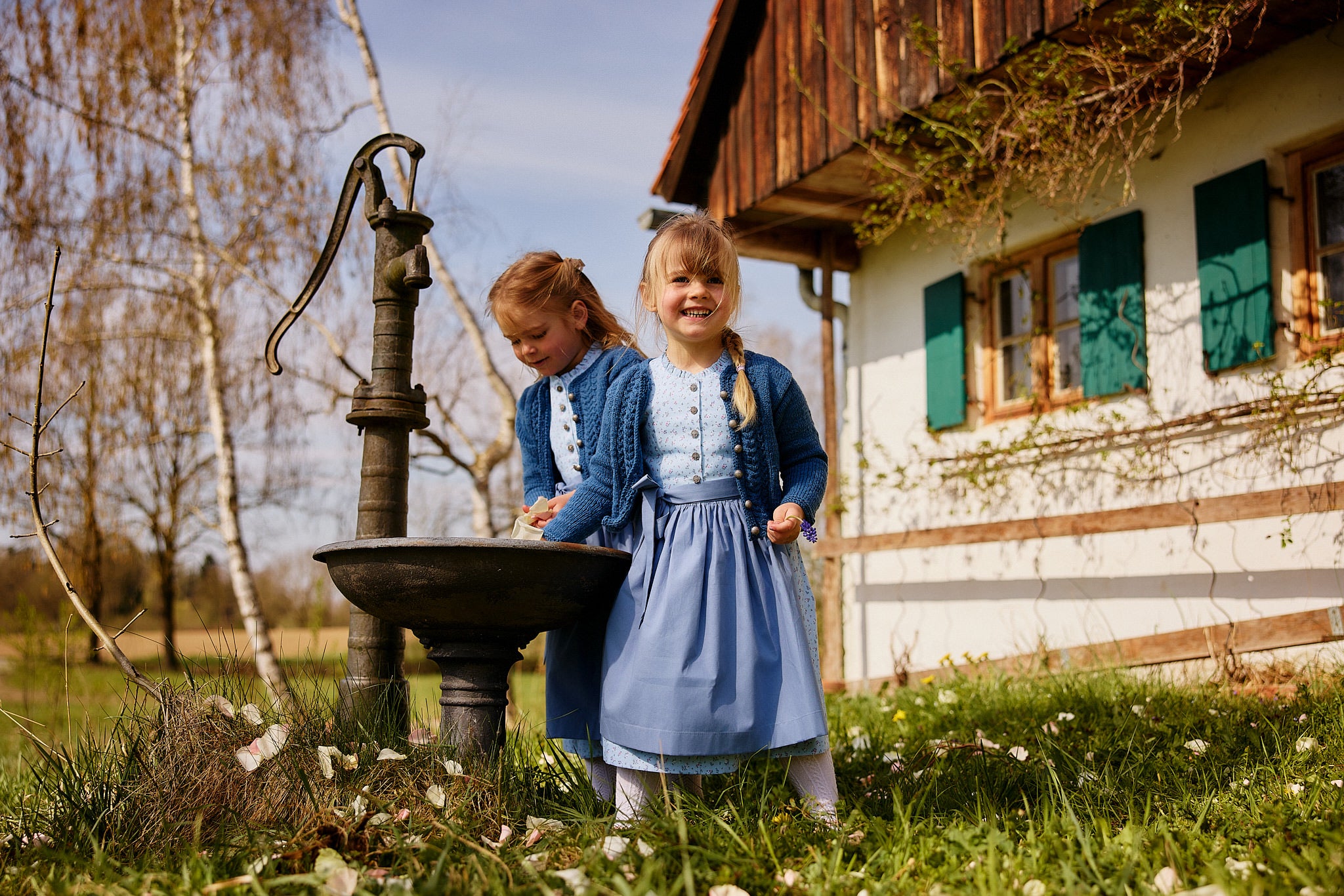 Kinderdirndl in hellblau aus Bio-Baumwolle mit blauer Schürze und blauer Strickjacke, beides handgemacht in Bayern, getragen von zwei Mädchen beim Spielen mit Brunnen
