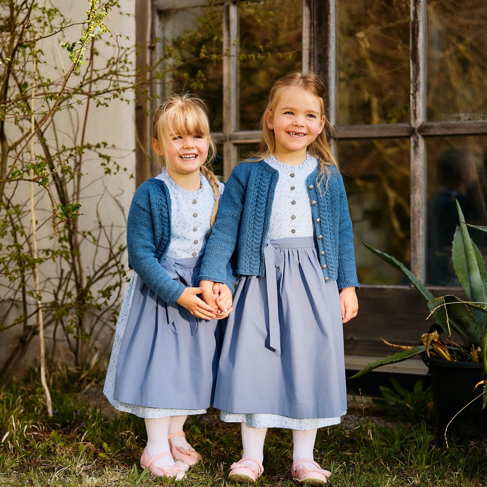 Kinderdirndl in hellblau mit passender blauer Kinderstrickjacke aus Bio-Baumwolle getragen von zwei Mädchen im Garten