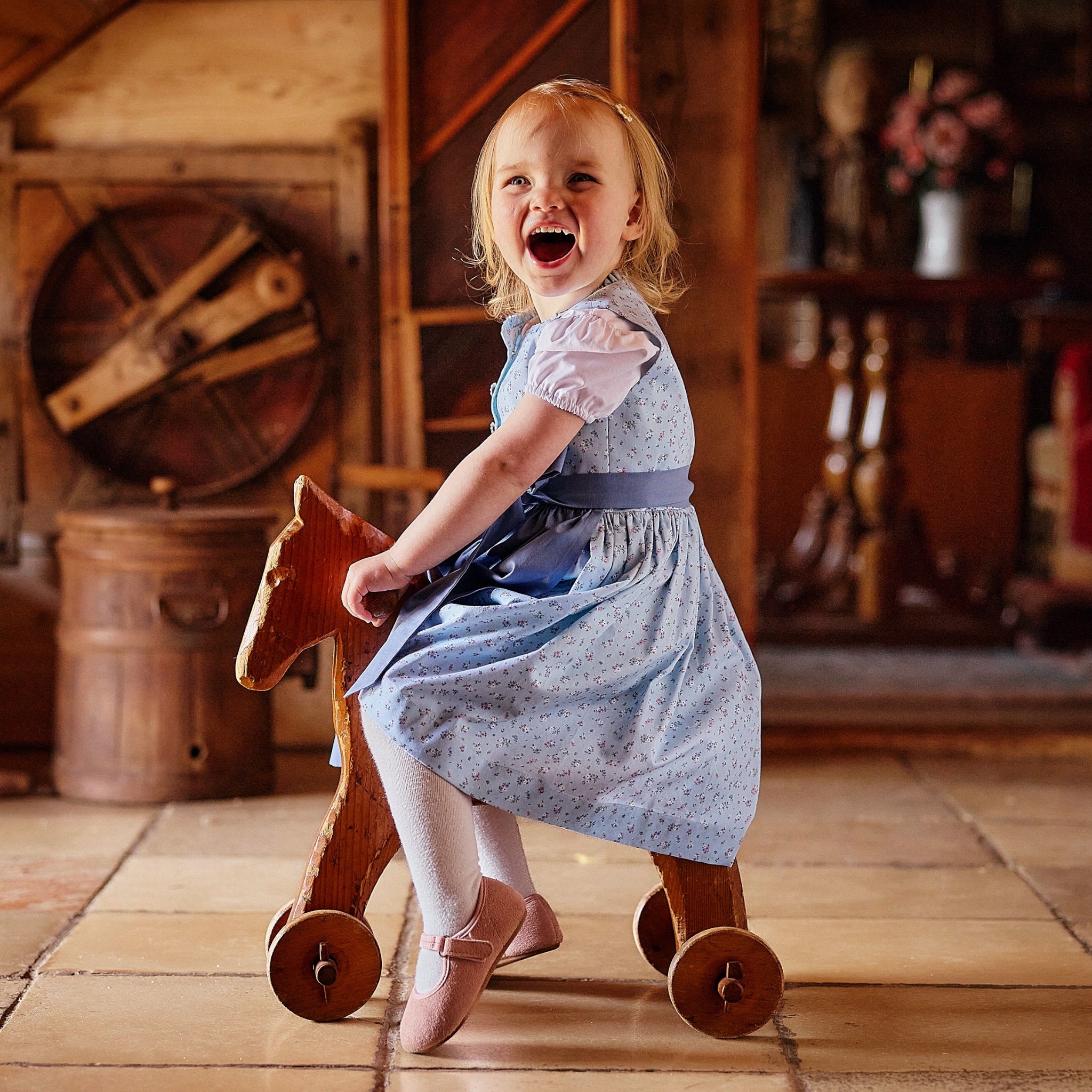 Kinderdirndl in hellblauem Allover Bio-Baumwollstoff mit Blumenmuster und blauer Schürz getragen von einem Kleinkind auf einem Schaukelpferd in einem alten Bauernhaus 