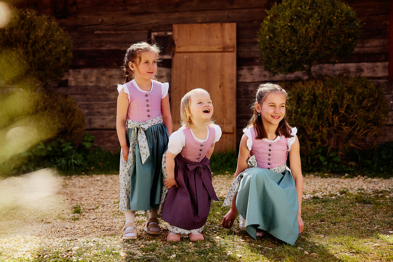 Kinderdirndl in flieder mit zwei unterschiedlichen Schüruzen in Salbei und Lila getragen von einem Kleinkind und zwei Mädchen